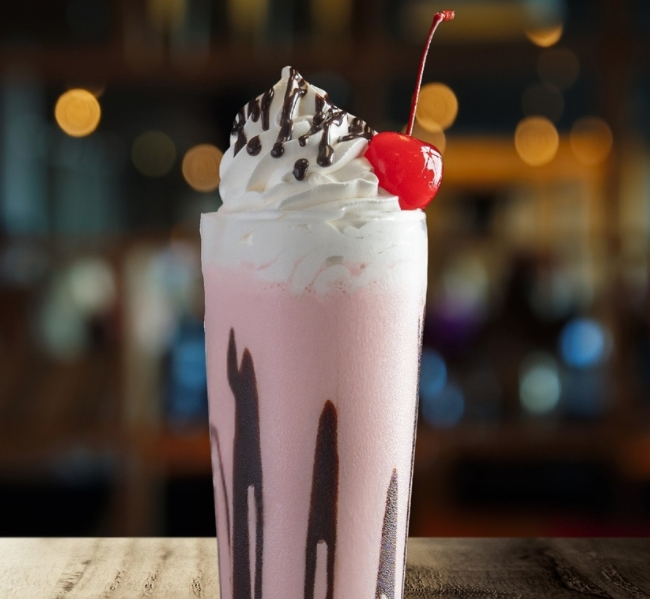 A pink milkshake from Gordon Ramsay Burger - Great Canadian Casino Vancouver, topped with whipped cream, chocolate drizzle, and a cherry, served in a tall glass on a wooden surface with warm blurred lights in the background.