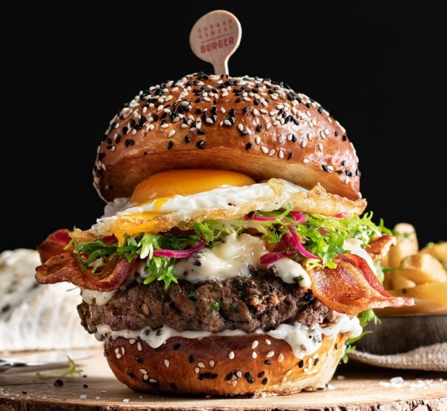 A gourmet burger on a sesame seed bun, topped with crispy bacon, a fried egg, melted cheese, fresh salad leaves, and creamy sauce—served on a wooden board with chips. Enjoy this delicious creation at Gordon Ramsay Burger - Great Canadian Casino Vancouver.