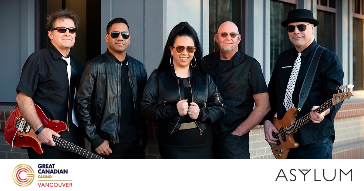 Five musicians stand outside a building, posing together. Three men wear dark sunglasses and black outfits, one holds a red guitar, another a bass. A woman stands at the centre, smiling, with a man wearing spectacles beside her.