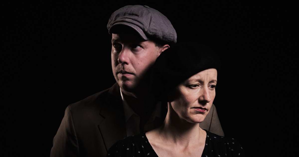 A man in a flat cap stands behind a woman in a black beret. Both look serious and pensive against a dark background, with soft lighting highlighting their faces.