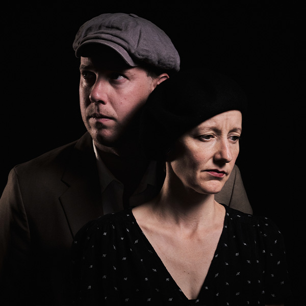 A man in a flat cap and a woman in a black beret stand close together against a dark background, both looking serious and thoughtful, with soft dramatic lighting highlighting their faces.