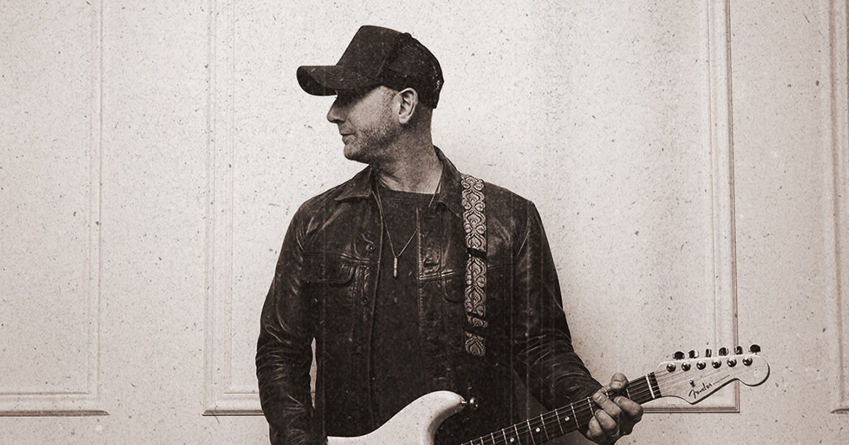A man in a dark jacket and baseball cap stands holding an electric guitar, looking to his left. The image has a vintage, grainy effect and a neutral background with simple panelling.