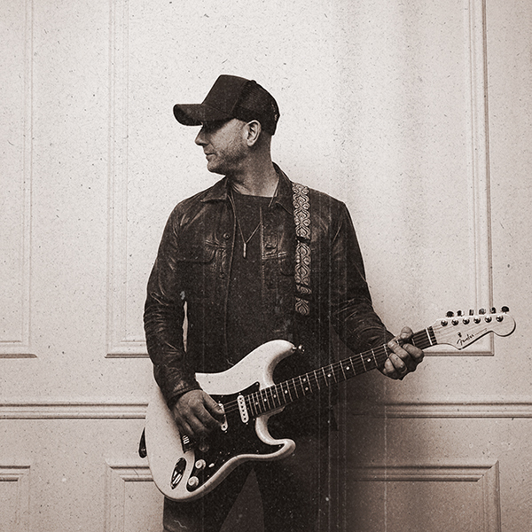 A man wearing a black cap and leather jacket stands against a white panelled wall, holding an electric guitar. He is looking to his left and appears to be ready to play. The image has a vintage, sepia-toned filter.