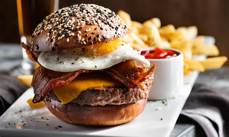 A close-up of a gourmet burger with bacon, cheddar cheese, and a fried egg on a seeded bun, served with chips and a small pot of tomato sauce on a white plate.