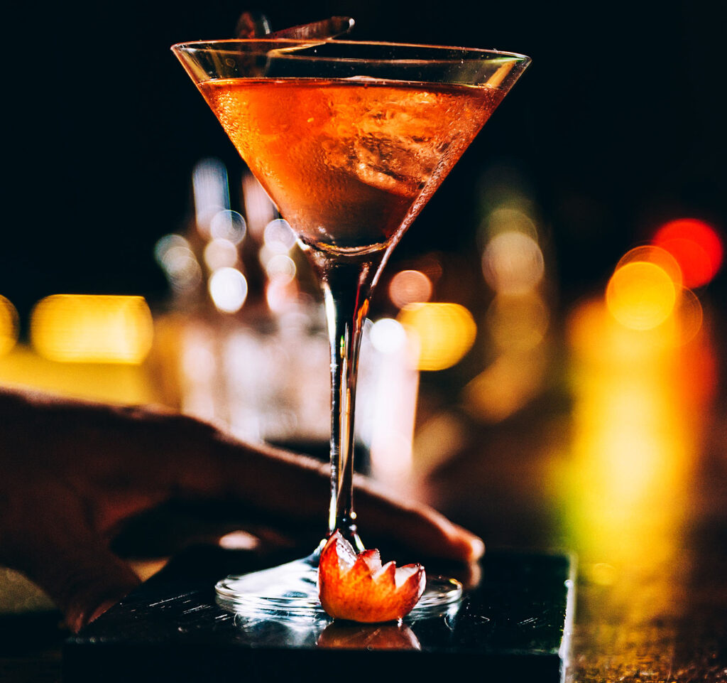 A hand holds a stemmed cocktail glass filled with an amber drink and ice, set on a dark surface with a sliced cherry garnish at Gordon Ramsay Burger - Great Canadian Casino Vancouver. Warm, blurred lights create a vibrant, inviting atmosphere.