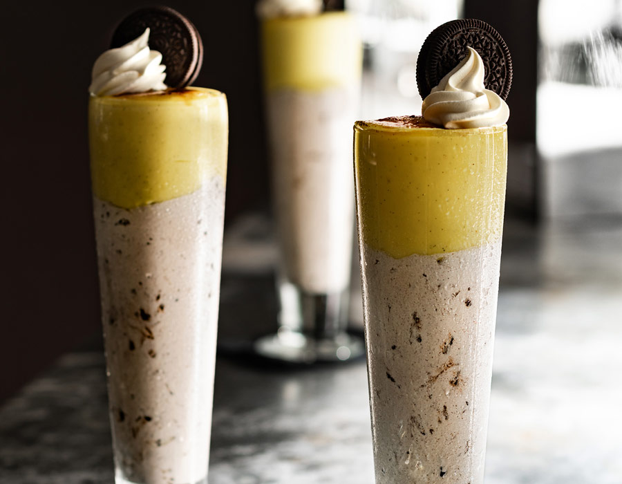 Three tall glasses filled with a layered dessert—biscuits and cream base, yellow custard top, each crowned with whipped cream and a chocolate biscuit. The display evokes the indulgence of sweet treats at Casino Vancouver on a marble surface.