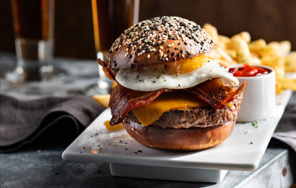 A hamburger with bacon, cheddar cheese, and a fried egg on a seeded bun is served on a white plate with chips and a cup of tomato sauce, with two drinks in the background.