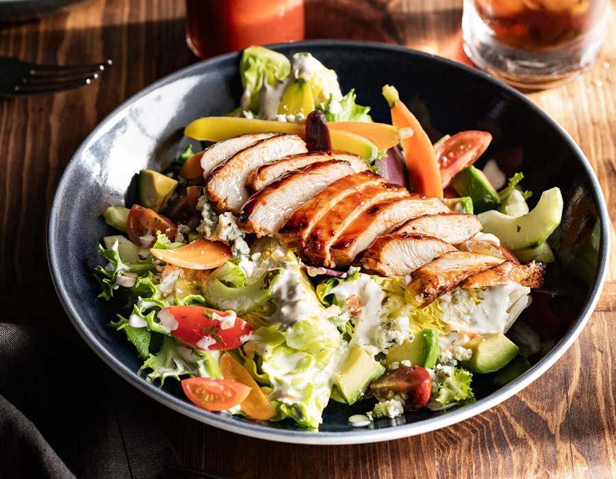 A bowl of fresh salad topped with grilled sliced chicken breast, surrounded by colourful vegetables and creamy dressing, is served on a wooden table—perfect for a healthy meal before a night out at Casino Vancouver.