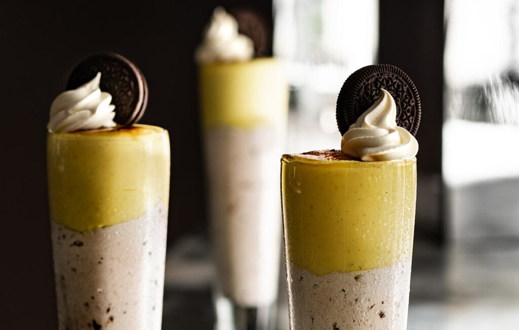 Three layered dessert glasses with cookies and cream mousse, yellow custard, topped with whipped cream and an Oreo biscuit, are artfully presented on a dark surface at Casino Vancouver, with a blurred background adding elegance.