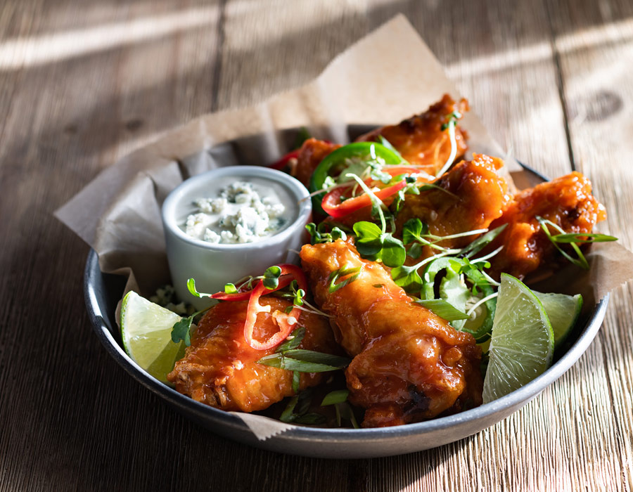 A metal bowl lined with baking parchment, filled with glazed chicken wings, lime wedges, fresh herbs, sliced red chilli, and a small pot of creamy dipping sauce on a rustic wooden table at Casino Vancouver.