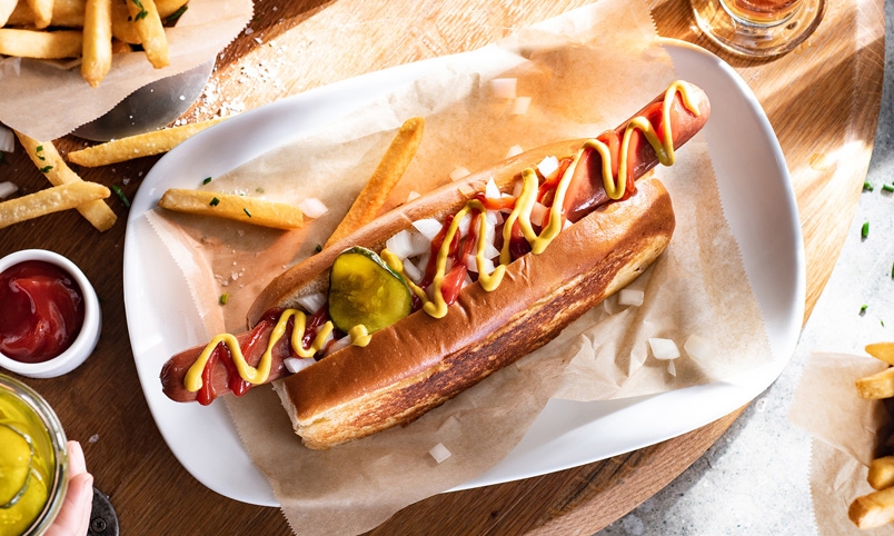 A hot dog topped with tomato sauce, mustard, chopped onions, and gherkins on a toasted bun, served on a white plate with a few chips. A dish of tomato sauce and more chips are nearby.