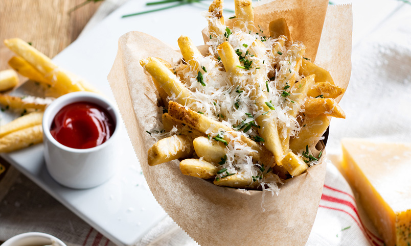 A cone of chips topped with grated cheese and chopped herbs sits on greaseproof paper, with a small pot of tomato sauce and a wedge of cheese in the background.