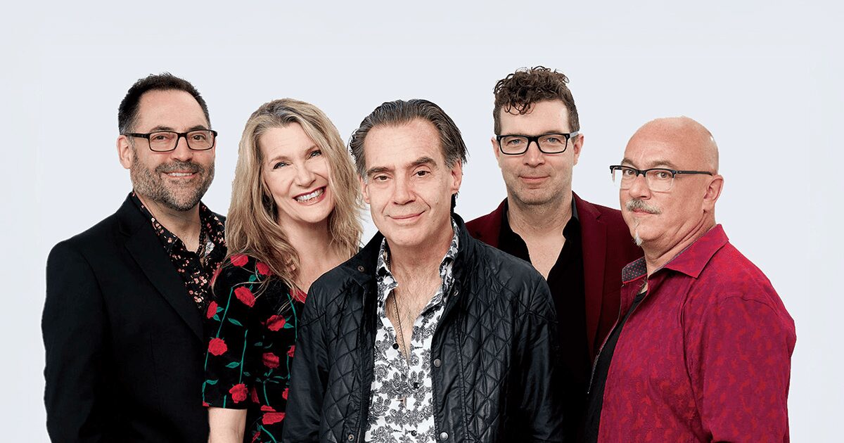 Five adults stand together against a plain light background, smiling or looking at the camera. They are dressed in casual and semi-formal clothing in dark and red tones.