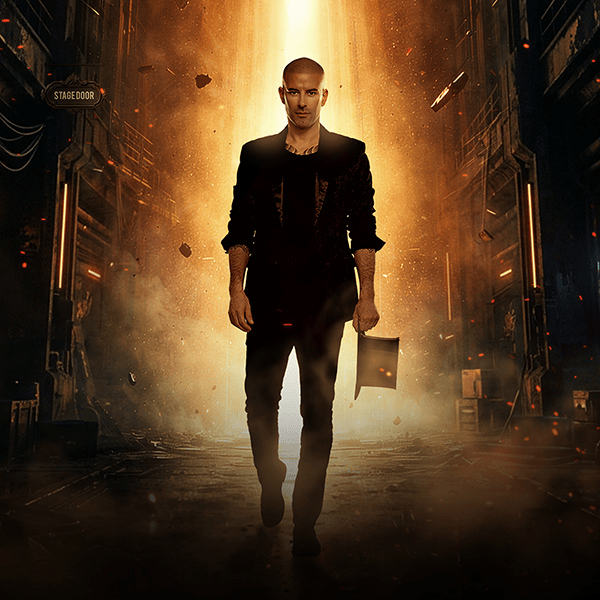 A bald man in a black suit walks confidently down a dramatic, glowing corridor with fiery orange light behind him, holding a folder. The setting appears industrial, with a "Stage Door" sign visible on the left.