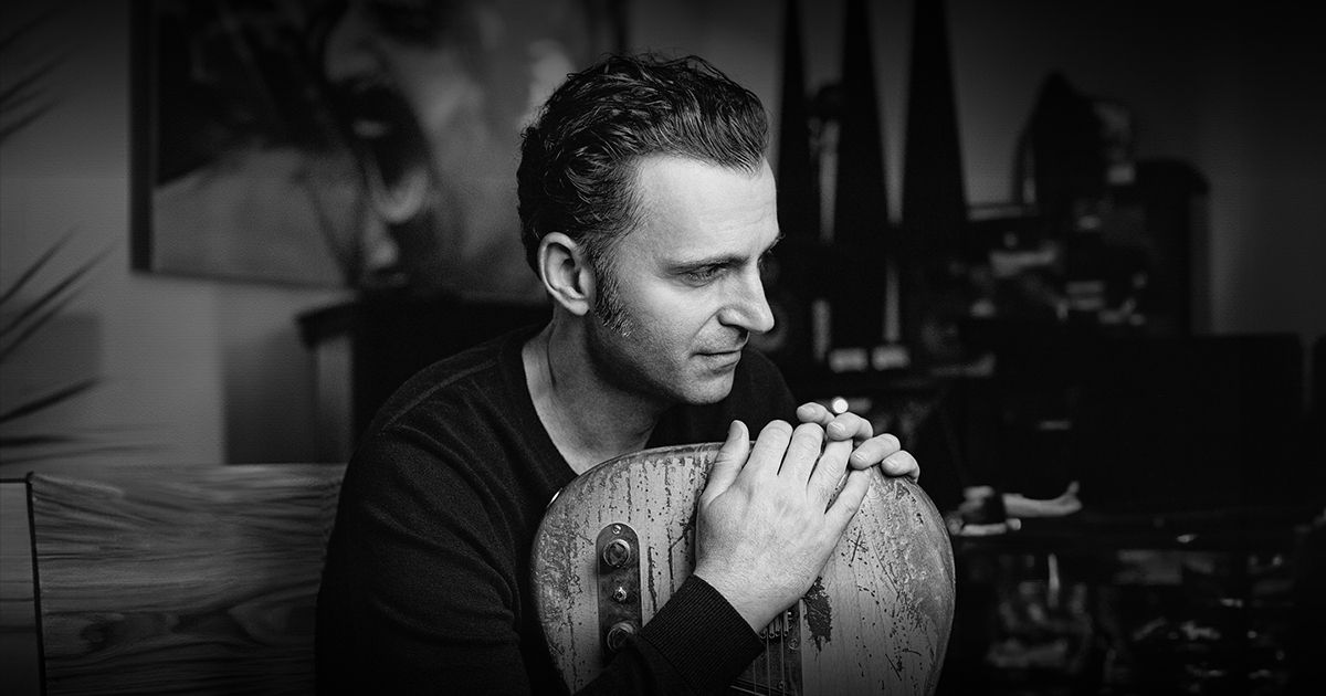 Black and white photo of a man with wavy hair sitting and leaning forward, resting his arms and chin on the body of an old, worn guitar, looking thoughtfully to the side.