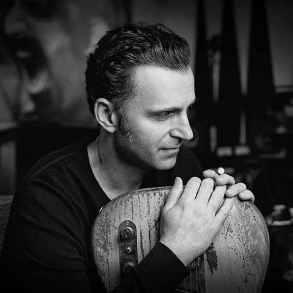 Black and white photo of a man with wavy hair sitting and holding a worn electric guitar, looking thoughtfully to the side. The background is blurred with abstract shapes and dark shadows.