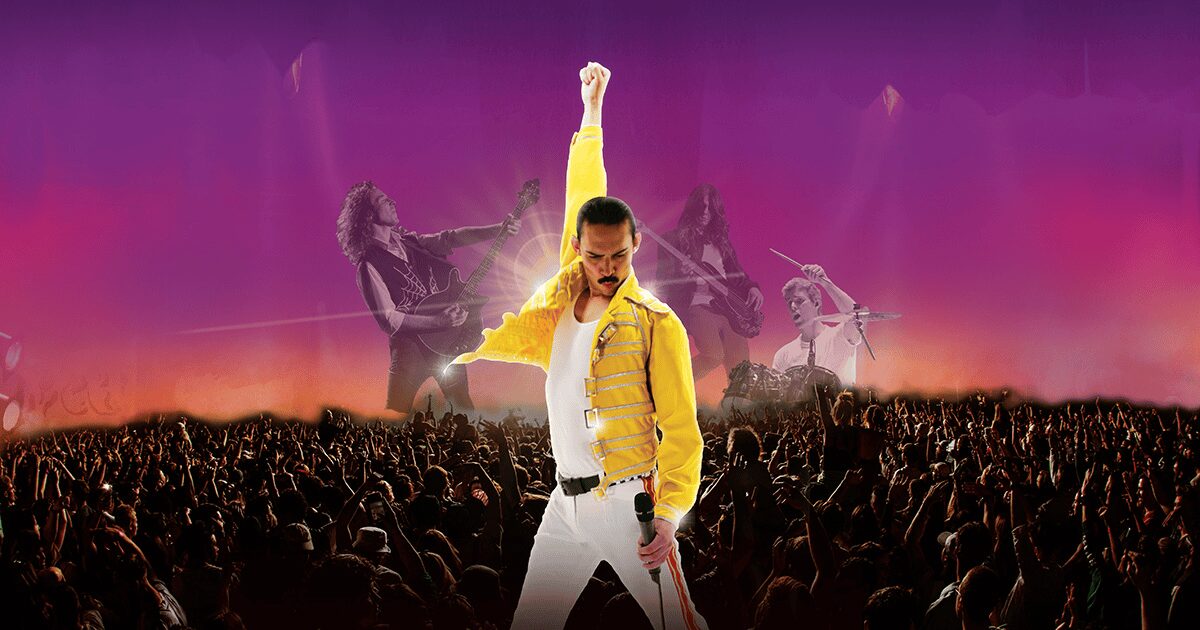 A performer in a yellow jacket stands on stage with one arm raised and holding a microphone, facing a cheering crowd, with band members and vibrant purple and orange lighting in the background.