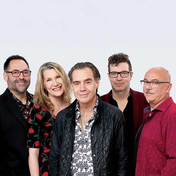 Five adults stand close together, smiling at the camera. They wear patterned or colourful shirts and jackets against a plain, light background. Four men and one woman are in the group.