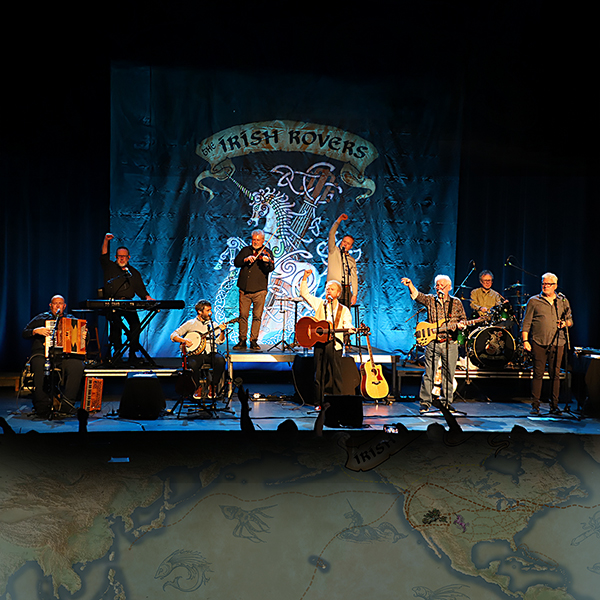 A band of eight musicians performs on stage with various instruments in front of a large backdrop reading "The Irish Rovers." The stage is lit in blue, and some band members raise their hands whilst playing.