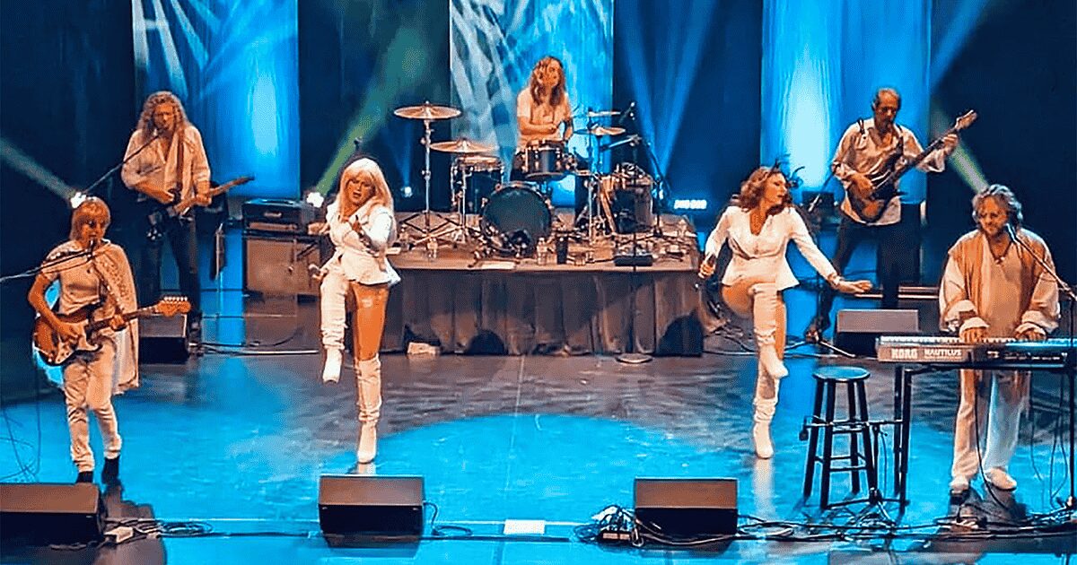 A band dressed in white costumes performs energetically on stage with blue lighting. Two women sing and dance at the front, whilst other members play guitar, keyboard, bass, and drums in the background.