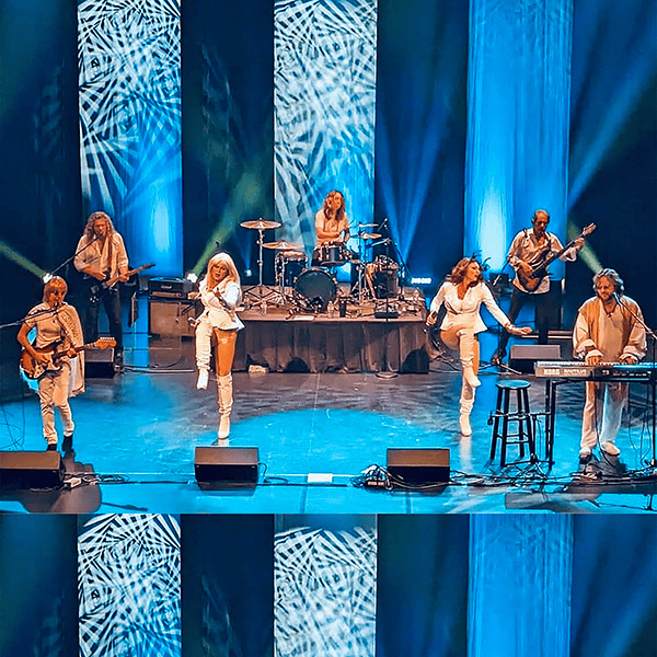 A band performs energetically on stage with blue lighting and patterned backdrops; two members are mid-jump while others play instruments including guitar, keyboard, and drums.
